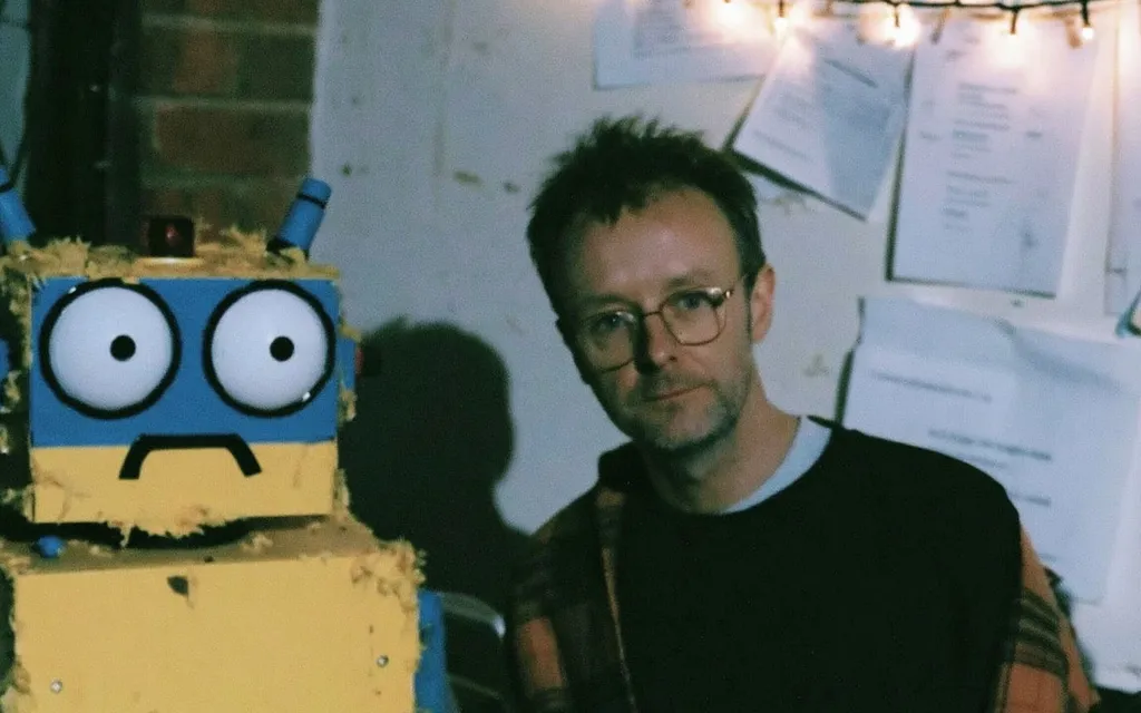 A person sitting beside a homemade cardboard robot with a worried face, a nod to AI agents that can act on command but can't yet answer the harder questions